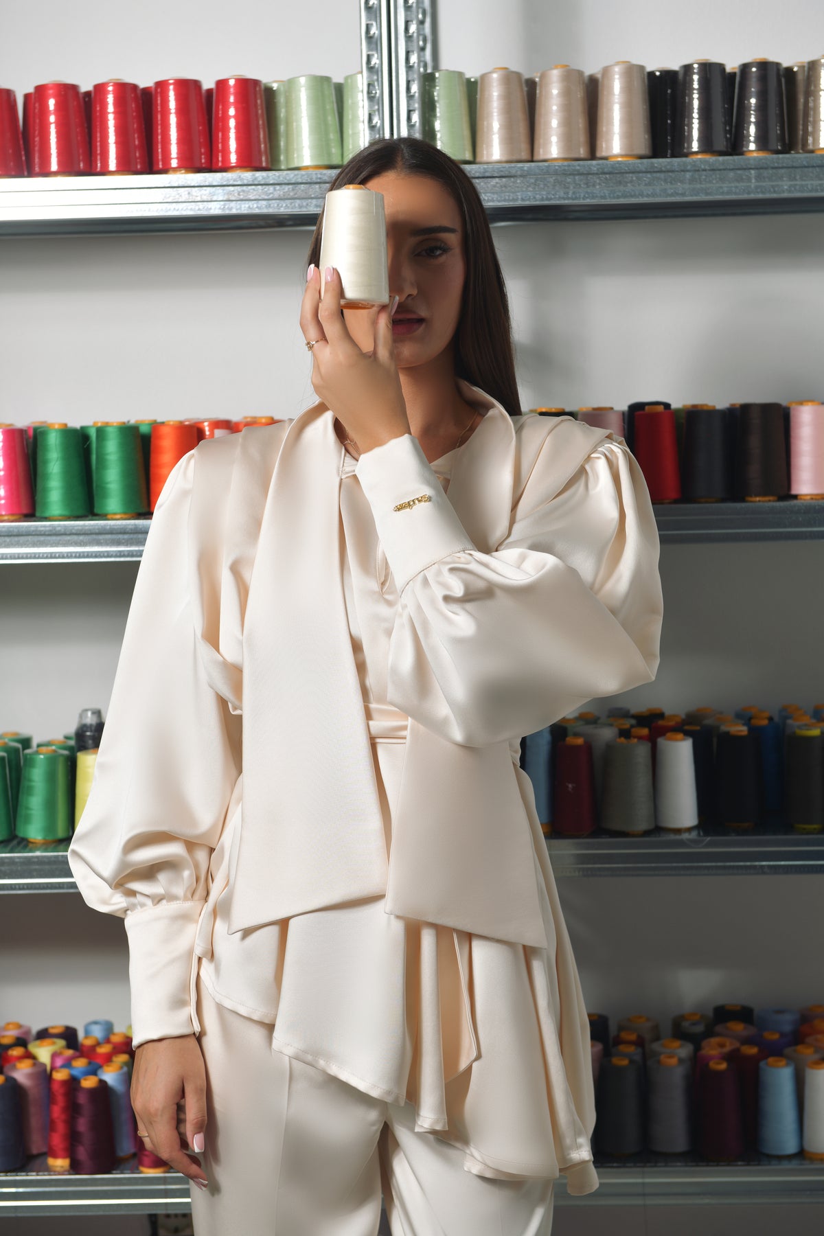 Model in a cream satin blouse and trousers in front of shelves of thread